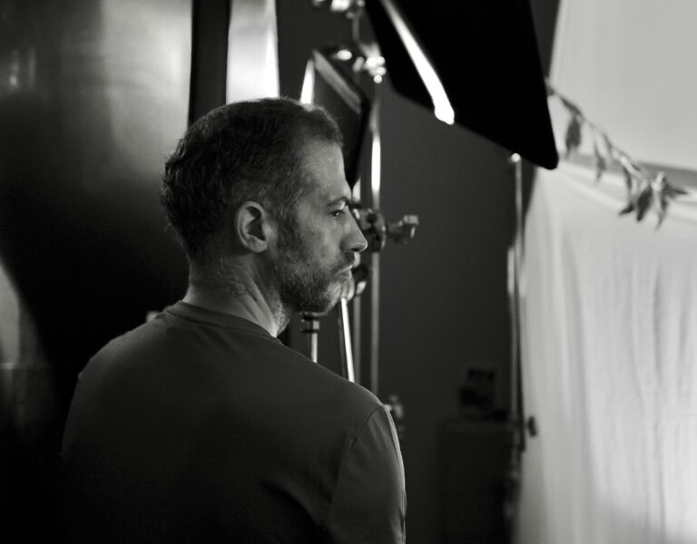 A man with short hair and a beard stands in a dimly lit studio, facing a camera and equipment. Light panels, stands, and a white backdrop are visible in the background.