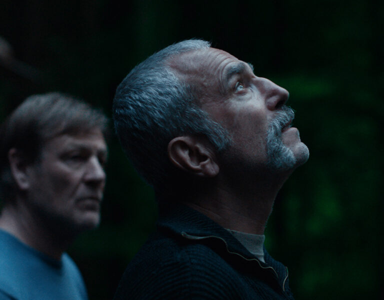 Two middle-aged men stand outside at dusk. The man in the foreground, Ben Fordesman, with short grey hair and a moustache looks upward, while the other man faces forward. A dark forest and anemone flowers are visible behind them.