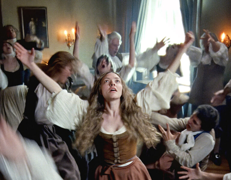 A young woman with long curly hair and a brown dress stands with arms raised in a crowded room as others around her gesture dramatically, in a scene with historical costumes and dim lighting at an EnergaCAMERIMAGE Film Festival Special Screening.