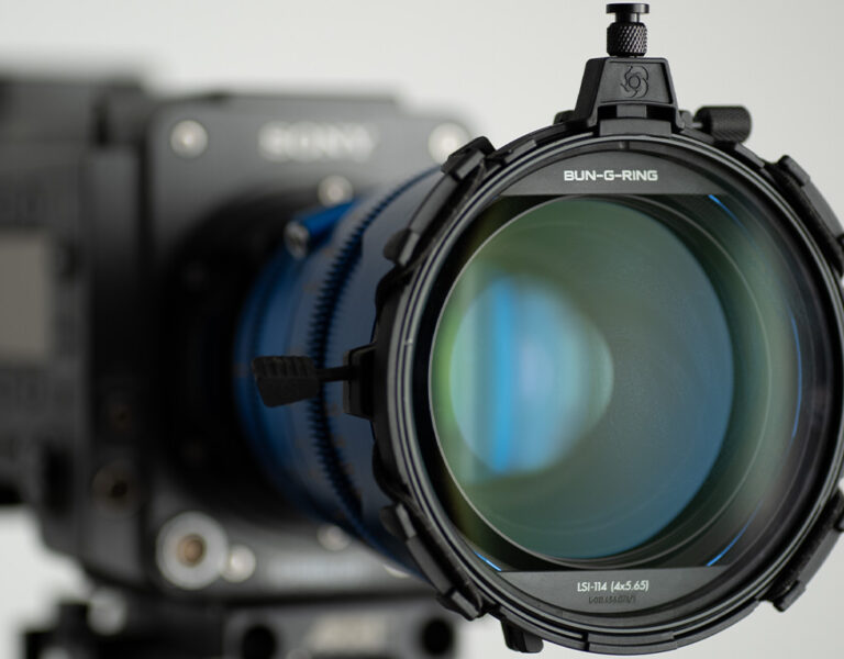 A close-up view of a professional video camera with a large lens and Bun-G-Ring Circular Glass Adapter attached, facing the viewer. The camera is mounted on a tripod against a neutral background.