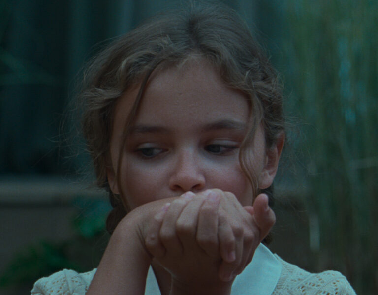 A young girl with wavy brown hair looks to her left whilst holding both hands up to her mouth. She is wearing a white top and is photographed in a natural, dimly lit setting.