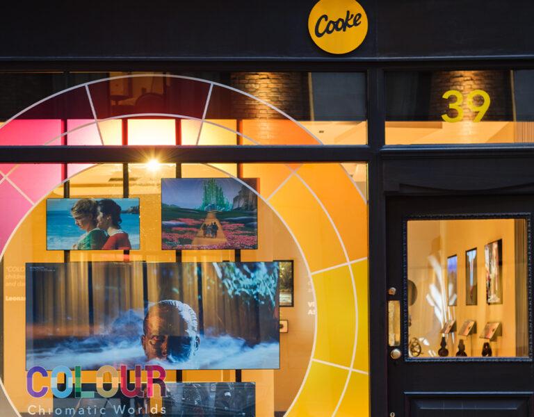 Shopfront with a large circular colour wheel graphic, “Cooke” logo above the door, and number 39 on the glass. Screens display various images for a Cooke Optics exhibition, with “COLOUR Chromatic Worlds” on the window.