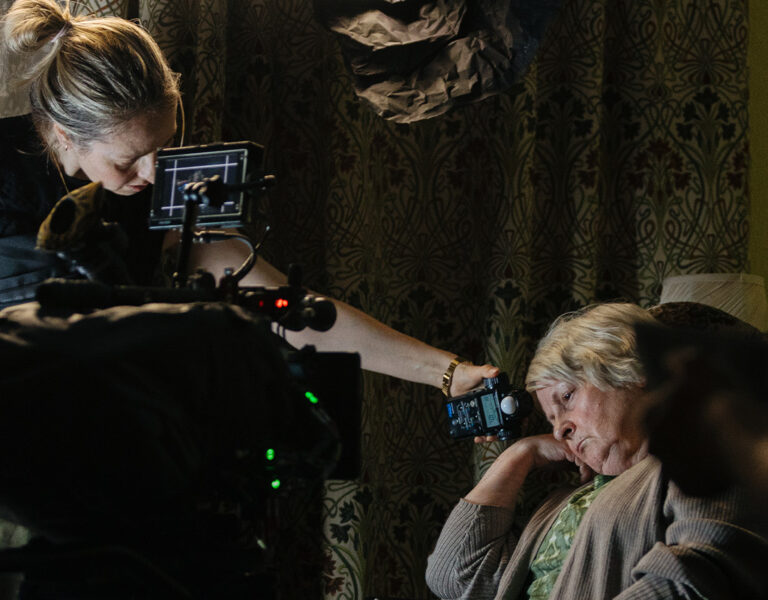 A woman adjusts a camera near an older woman seated in a chair with her eyes closed. Studio equipment and patterned curtains are visible in the background, suggesting a Dragonfly production set with cinematographer Vanessa Whyte BSC.