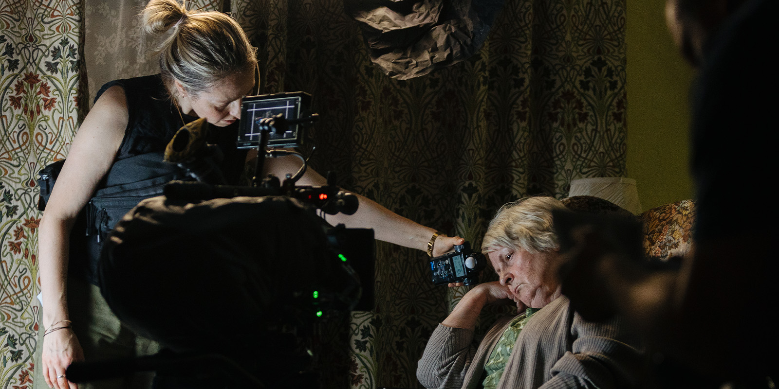 A woman adjusts a camera near an older woman seated in a chair with her eyes closed. Studio equipment and patterned curtains are visible in the background, suggesting a Dragonfly production set with cinematographer Vanessa Whyte BSC.