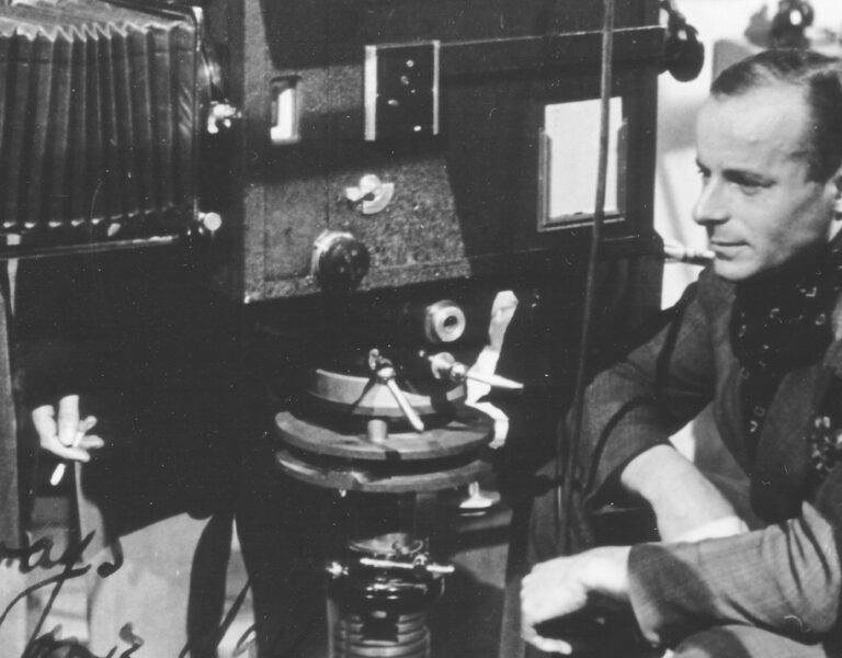 A man in a suit sits beside a large, old-fashioned film camera and equipment. He appears focused, with one hand resting on his knee. The black-and-white scene suggests a BSC Heritage Series set, evoking Erwin Hillier’s era of classic cinematography.