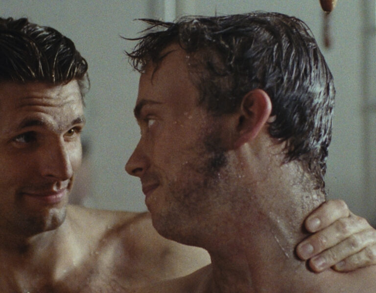 Two bare-chested men stand close together in a shower, gazing at each other with playful smiles. Water droplets glisten on their skin, capturing an intimate moment reminiscent of "Every Time I See a Cock I Go Funny" by Catharina Scarpellini.