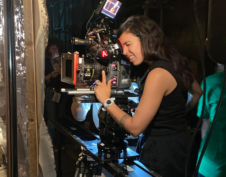 A woman operates a professional movie camera on a set, adjusting controls and looking through the viewfinder. Crew members and film equipment surround the dimly lit area, reminiscent of scenes from Every Time I See a Cock I Go Funny.