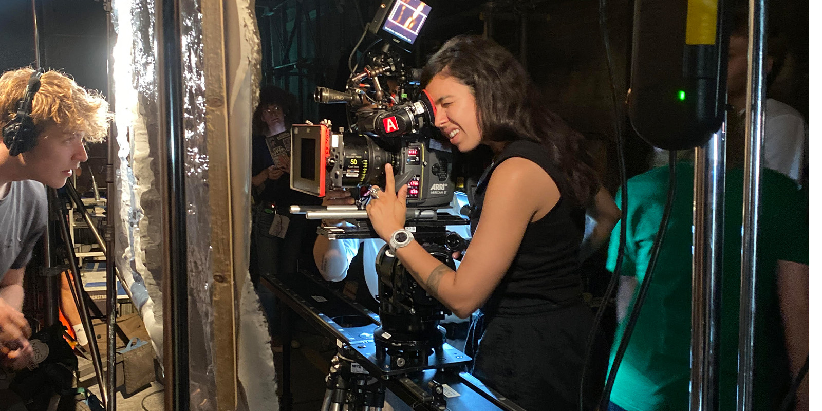 A woman operates a professional movie camera on a set, adjusting controls and looking through the viewfinder. Crew members and film equipment surround the dimly lit area, reminiscent of scenes from Every Time I See a Cock I Go Funny.