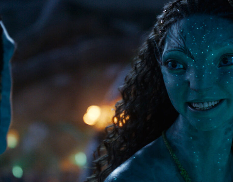 A blue-skinned, humanoid character with bioluminescent spots, yellow eyes, and curly hair smiles whilst facing another similar figure in a scene reminiscent of Russell Carpenter’s work on Avatar: Fire and Ash, softly lit with warm, out-of-focus lights.