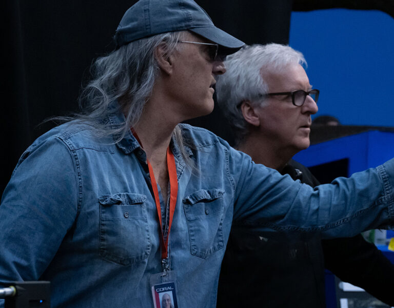 Two men stand next to each other indoors, one wearing a denim shirt, cap, and lanyard—resembling Russell Carpenter—whilst the other wears dark clothing and glasses. Both look at a monitor, possibly reviewing Avatar: Fire and Ash footage amidst blue and black elements.