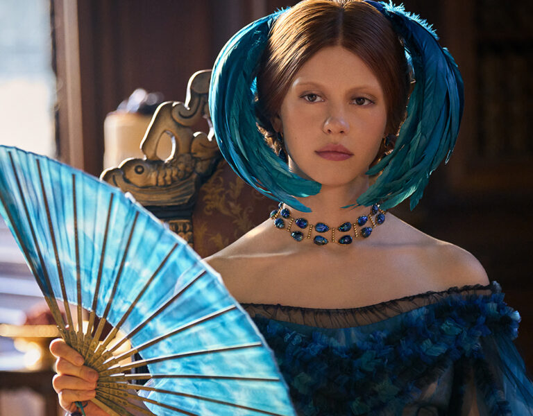 Mia Goth in a blue dress holding a fan