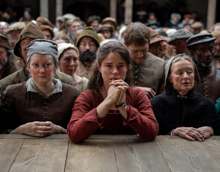 A large crowd in old-fashioned clothing stands closely behind a wooden barrier, their faces serious and sombre. At the centre, a young woman clasps her hands—a scene reminiscent of Hamnet, captured with Łukasz Żal’s evocative PSC cinematography.