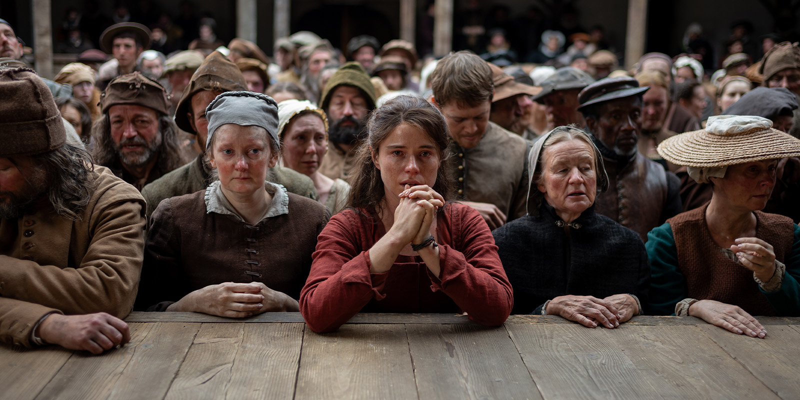 A large crowd in old-fashioned clothing stands closely behind a wooden barrier, their faces serious and sombre. At the centre, a young woman clasps her hands—a scene reminiscent of Hamnet, captured with Łukasz Żal’s evocative PSC cinematography.