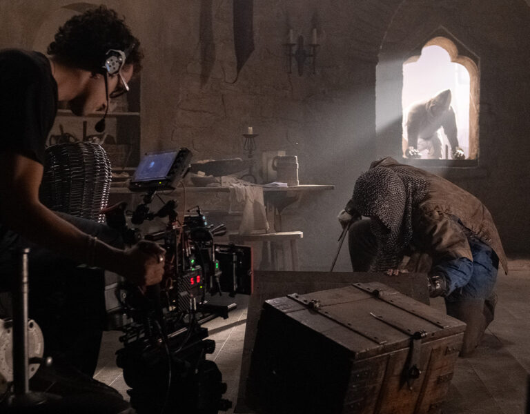 A film crew member operates a camera inside a dimly lit room with medieval decor, capturing an actor in costume kneeling by a wooden chest—reminiscent of a scene from Hand to God—whilst another figure stands framed in sunlight, Dylan Bruce-style.