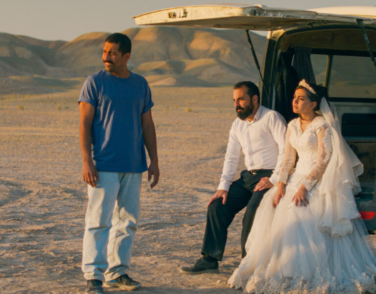 A bride in a white gown and a groom in a white shirt sit at the open back of a white van in a desert landscape. Another man, possibly Amin Jafari from "It Was Just an Accident," stands nearby, gazing into the distance.