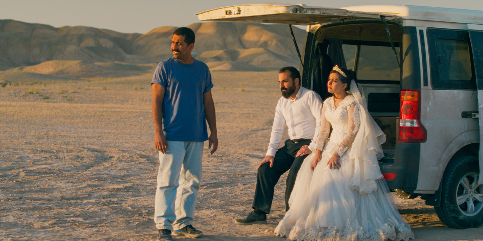 A bride in a white gown and a groom in a white shirt sit at the open back of a white van in a desert landscape. Another man, possibly Amin Jafari from "It Was Just an Accident," stands nearby, gazing into the distance.