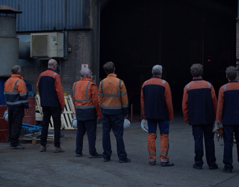 Seven people wearing orange and navy work uniforms stand with their backs to the camera, facing the entrance of an industrial building. The area has industrial equipment, signs, and some scattered materials.