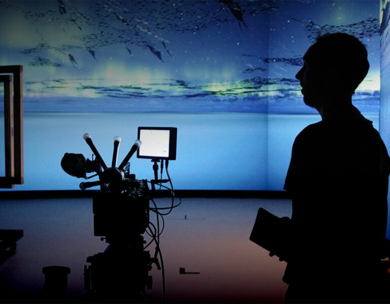 A person stands near a camera set-up in a studio with blue digital visuals projected on the walls.