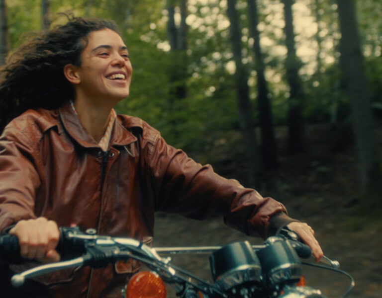 A person with long curly hair wearing a brown leather jacket rides a motorbike through a wooded area, smiling whilst looking ahead.