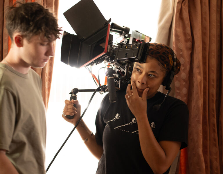 Adenike Oke, in a black shirt, holds a professional video camera on her shoulder whilst standing next to another focussed individual. Curtains and soft lighting set the scene as they capture footage, keeping their creative body count high.
