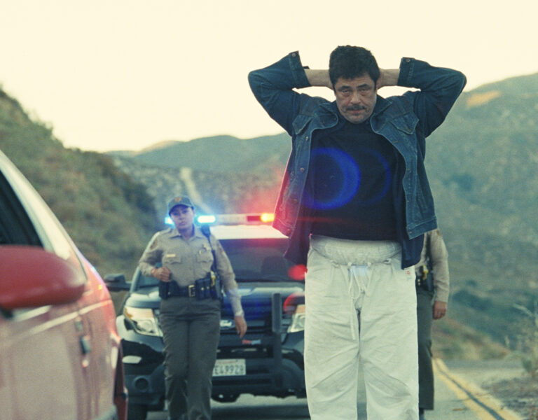 A man with his hands on his head stands on a road in front of a police car with flashing lights. Two officers approach him, while a red car and mountains are visible in the background.