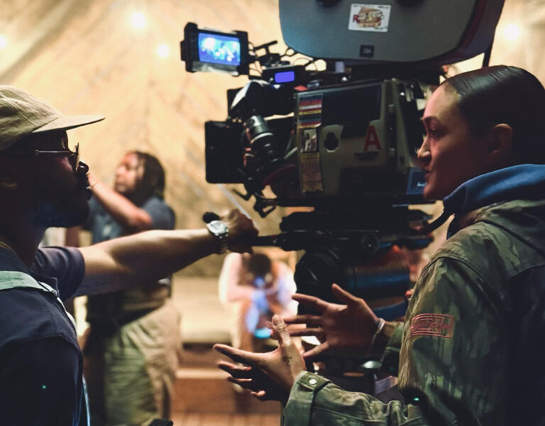 Two people stand in front of a professional film camera, engaged in discussion about film formats on a set with blurred crew and a warm, wooden backdrop.