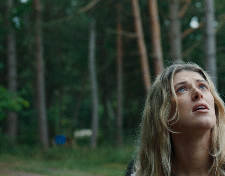 A woman with long blonde hair looks upwards with a concerned expression whilst standing outdoors in a wooded area. Tall trees and part of a vehicle are visible in the background.