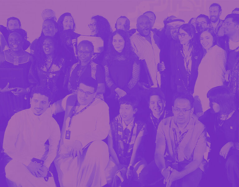 A large group of people pose together for a photo, some standing and some kneeling at the front, all facing the camera. The image has a purple colour overlay and was taken at the Red Sea International Film Festival's Red Sea Souk Project Market.