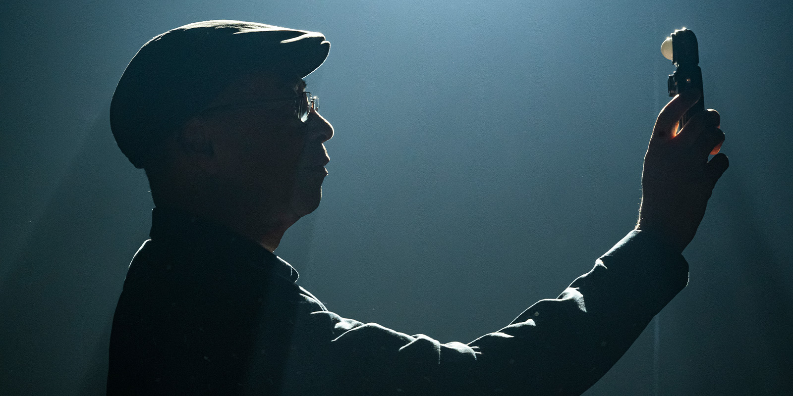A person wearing a cap holds up a handheld device in front of their face. The photo, reminiscent of cinematographer Remi Adefarasin, OBE's style, is taken in low light, creating a dramatic silhouette effect.
