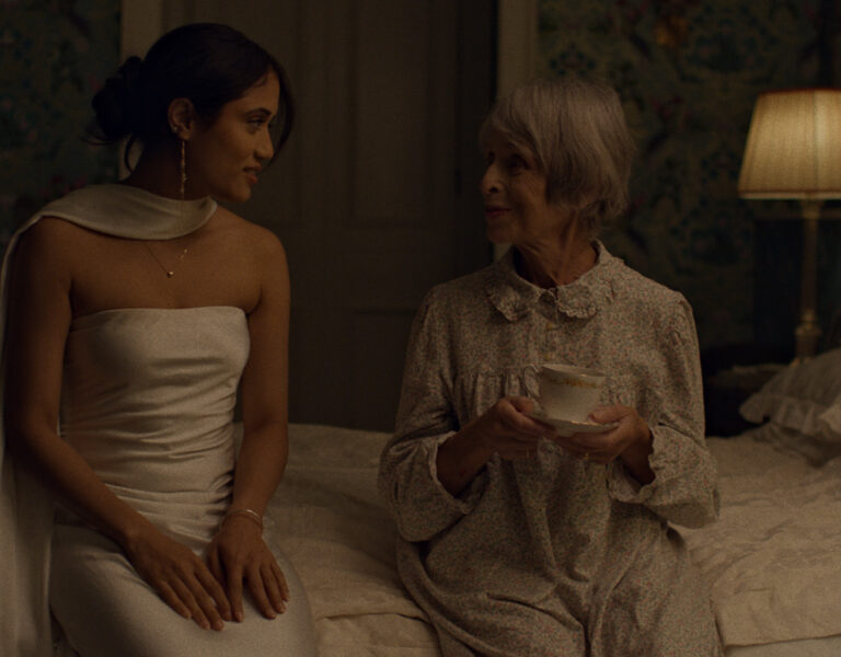 A young woman in a white dress sits on the edge of a bed next to another person, who is in pyjamas holding a teacup. They face each other and appear to be having a conversation in a dimly lit bedroom.