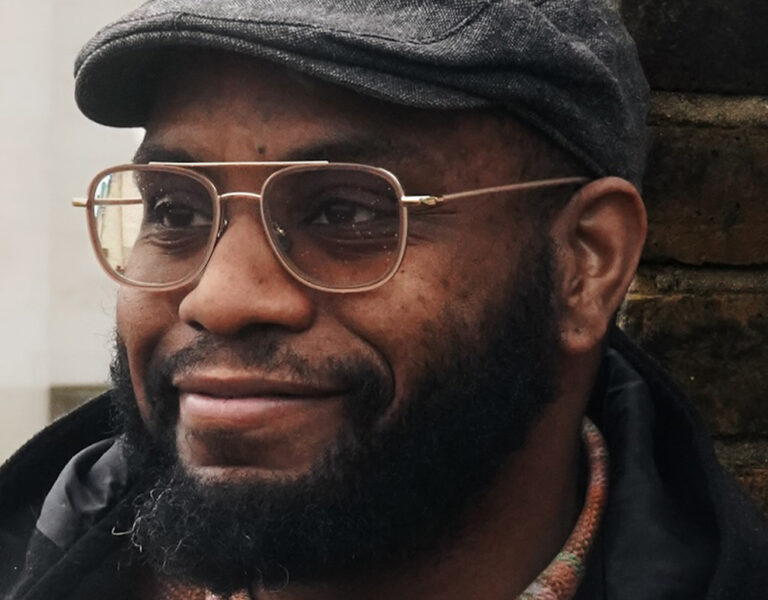 Stephen Huggy Henry, head of 3D at UNIT, stands in front of a brick wall wearing a flat cap and dark jacket. With his beard and glasses, he looks slightly to the side and smiles.