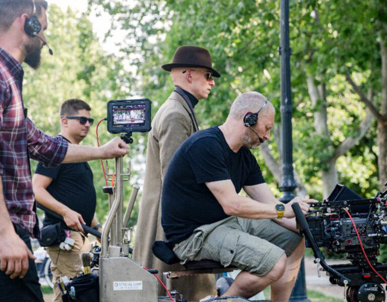 A film crew works outdoors with a camera set-up on a dolly, as Christopher Ross BSC operates the camera while others assist, wearing headsets. A person in a brown coat and hat walks nearby. Trees and park greenery are visible in the background.