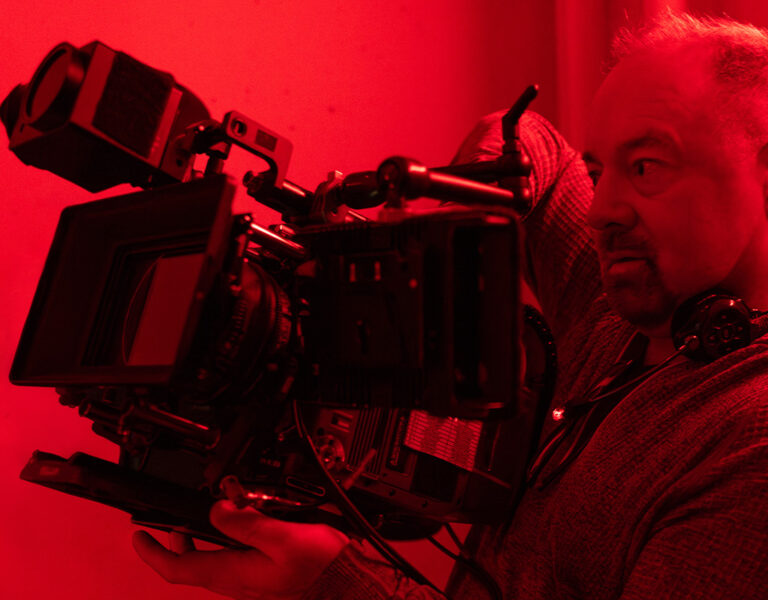 A person holding a large professional video camera on their shoulder, standing against a red-lit background, appears focused on filming in a style reminiscent of Jeff Cronenweth’s cinematography for Tron: Ares.