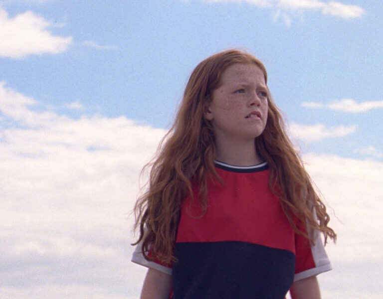 A young person with long red hair stands outdoors under a partly cloudy sky, wearing a red and black shirt and looking off into the distance with a serious expression.