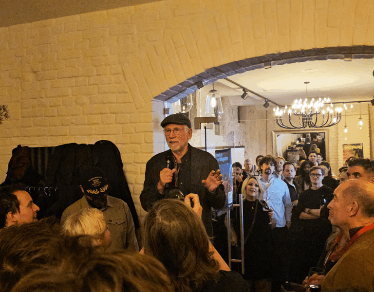 A man stands and speaks into a microphone to a crowded room in a warmly lit space with cream brick walls, a chandelier, and decorative elements. The scene captures 2025-inspired cinematography as most people focus on the speaker.